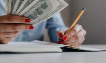 A woman's hands holding and counting cash, while writing in a notebook for blog post make an extra $500 a month.