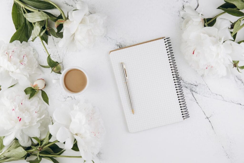 A pen, notebook, coffee, and flowers on a marble countertop.
