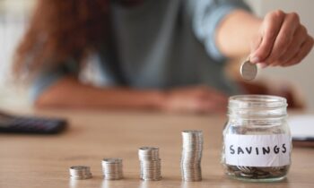 A woman counting coins in piles on a desk and then putting them into a jar labeled "savings" for the article clever ways to save money
