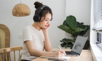 A woman sitting at her kitchen table, wearing headphones and working on a laptop.