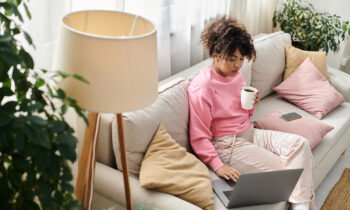 A young woman sitting on her sofa drinking coffee searching on her laptop for online work at home courses