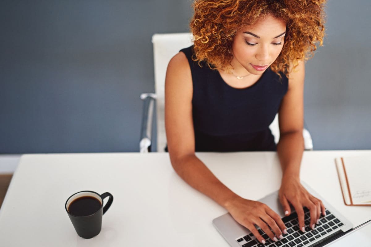 Overhead view of a woman working at her home office desk, drinking coffee and typing on a keyboard for blog post earn what you deserve.