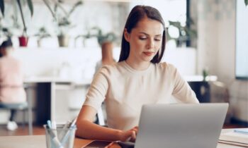 A woman sitting at a table working in a cafe on her laptop, responding to a rejection email.